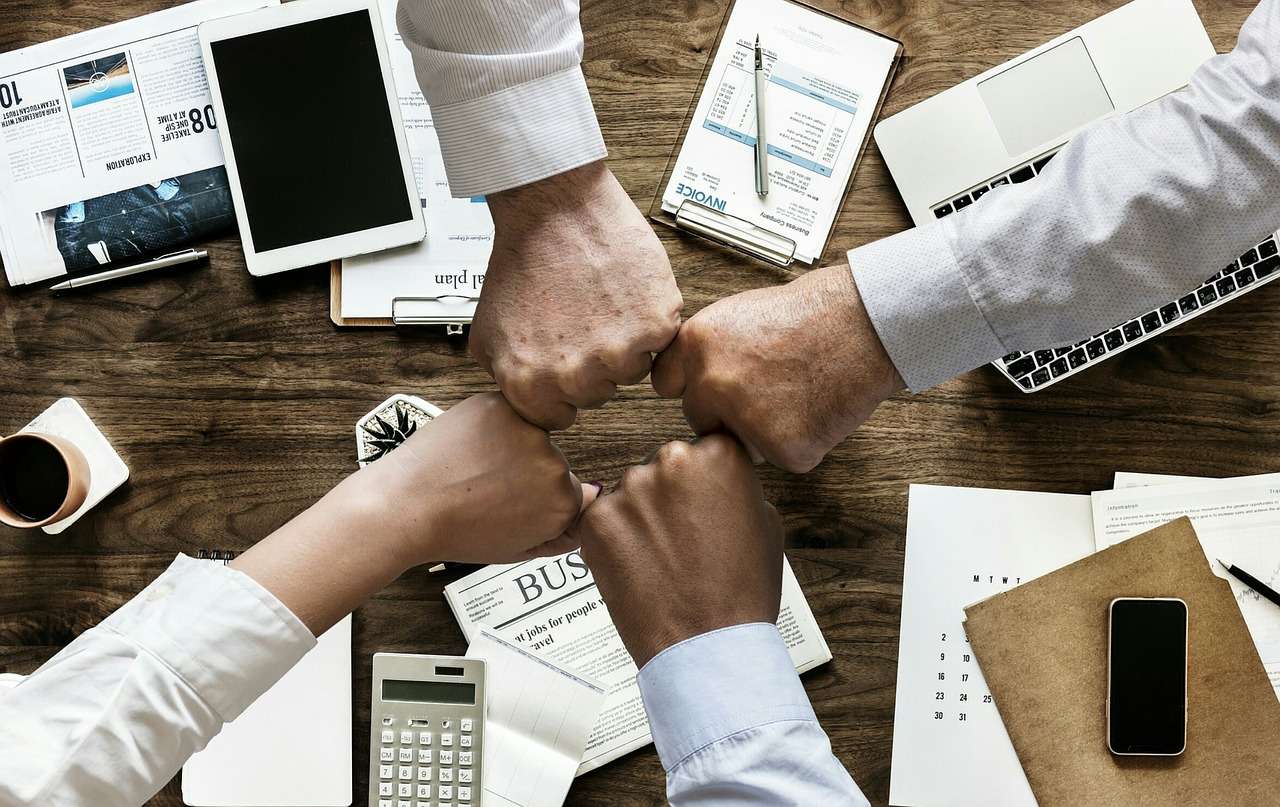 Forschungsteam arbeitet gemeinsam an Publikation, vier Hände stoßen zusammen über Schreibtisch mit Laptop, Tablet und Dokumenten